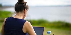 Photo of a woman studying outside with a laptop overlooking a lake  